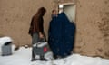 A woman in a full black chador-like veil holding a baby, enters a mud-walled compound with a man following holding a box