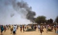 A large crowd of Africans running across a desert-like landscape as black smoke billows from a compound in the background
