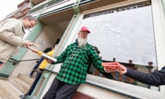 Dutch angle shot of a man in a green and black checked shirt and a red cap standing in front of a bookstore receiving a book from someone to his right and passing a book to someone on his left.