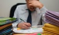 A stressed teacher marking work at a desk behind two piles of educational books