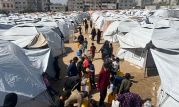 People with yellow water containers stand surrounded by white tents