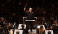 A conductor wields a baton in front of an orchestra looking at sheet music
