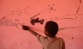 A young African boy points to a child's drawings on a wall of pickup trucks mounted with guns
