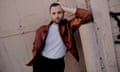Penn Badgley in brown leather jacket, white T-shirt and black trousers leaning against an industrial pillar
