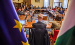People sit around tables arranged in a large rectangle with the EU and Hungarian flags in the foreground