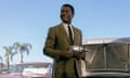 In a suit, he stands in a used car lot holding a drill